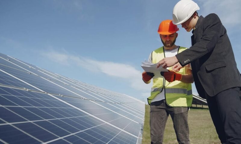 Low angle of young inspector and foreman in hardhats checking documentation against modern solar panels in field