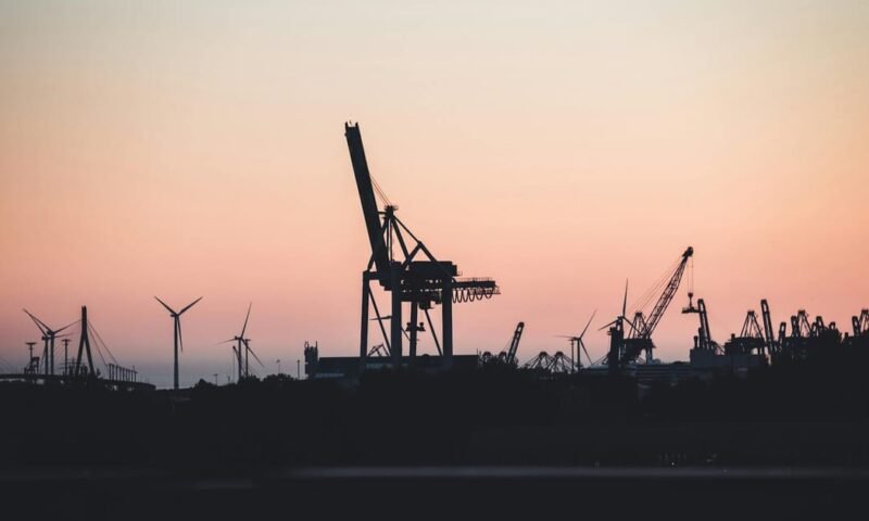 Silhouetted cranes and wind turbines at the port of Hamburg during sunset, highlighting industrial beauty.