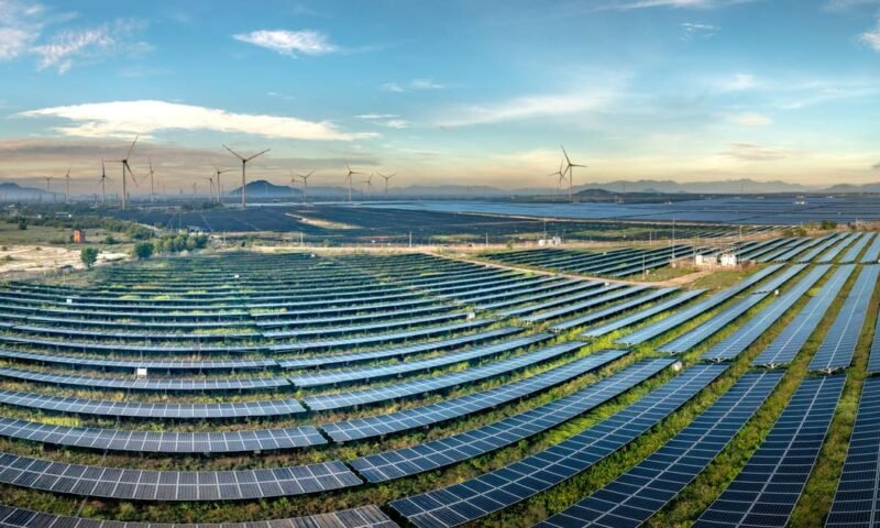 Expansive solar farm with wind turbines in the background under a clear blue sky.